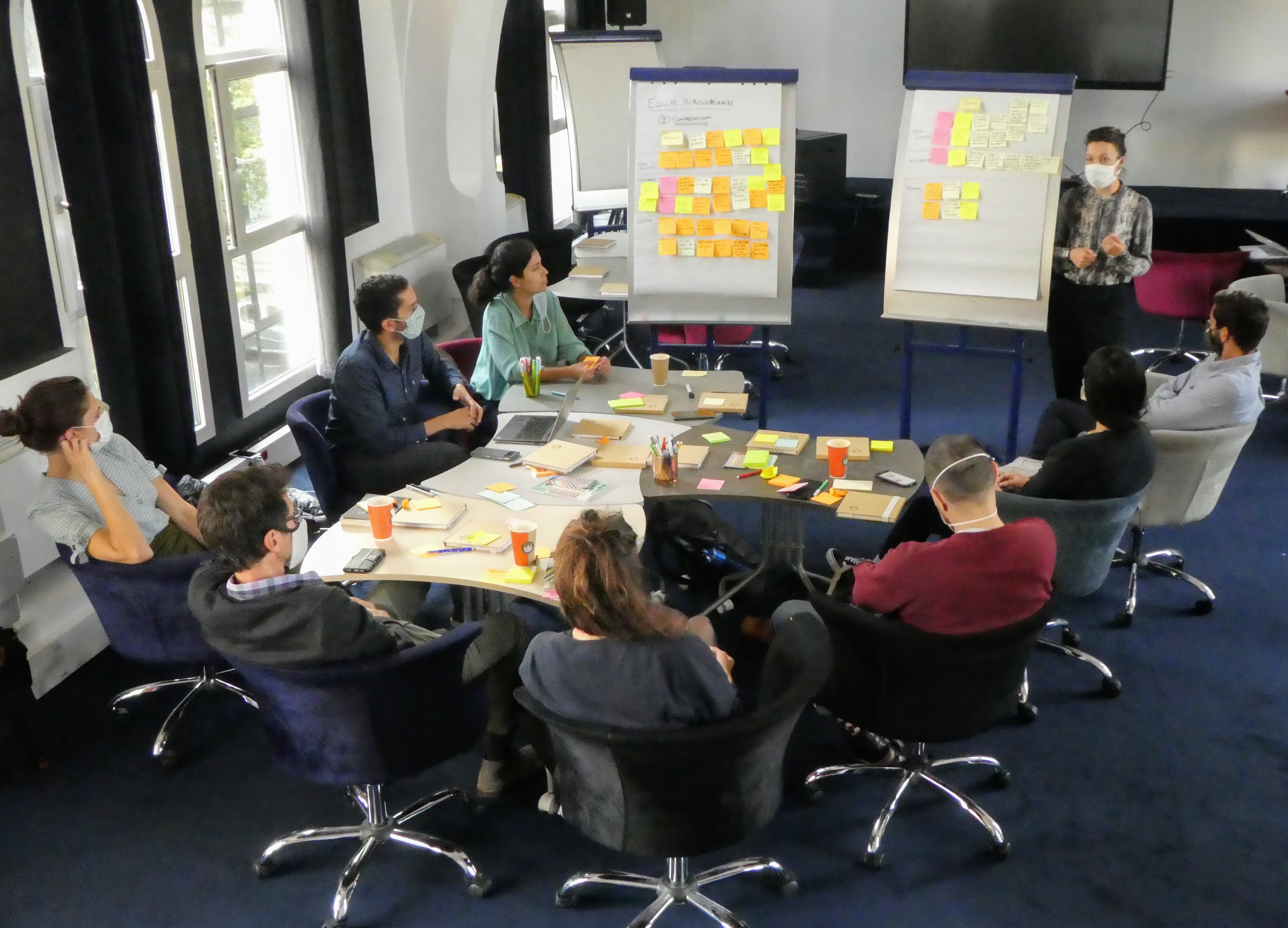 Huit personnes sont assises autour d'une table. Une personne présente. Les huit personnes font face à deux tableaux blancs, sur lesquels ont été collés des post-its.