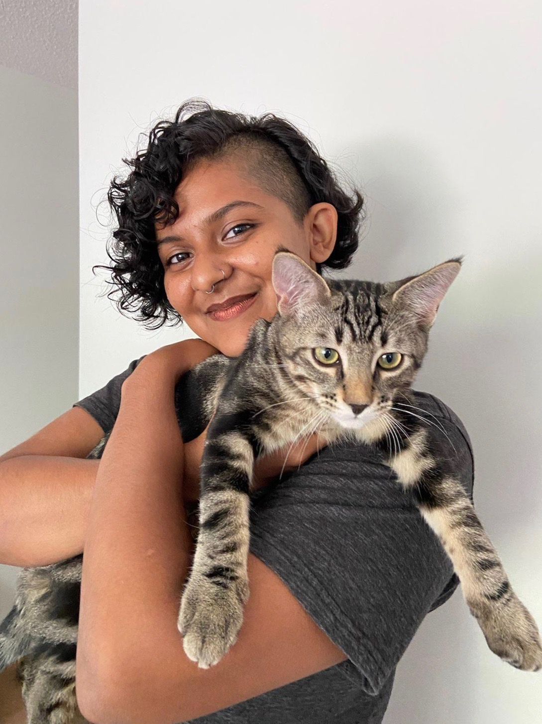 Vasundhara smiling at the camera holding Laszlo, a striped 5-month old kitten. Laszlo is a striped tabby x shorthair kitten and his owners Danica and Seb suspect that he probably also has some Bengal cat in him. Vasundhara is brown-skinned and has short curly black hair with a side buzz. Xe has a nose ring and a septum ring and is wearing a grey t-shirt. Laszlo's paws hang over xyr left arm and he looks very soft.