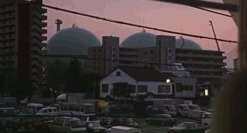 An animated gif backwards tracking shot from the movie 'Laundry' showing a woman staring out of her window at three large spherical gas tanks in the distance.