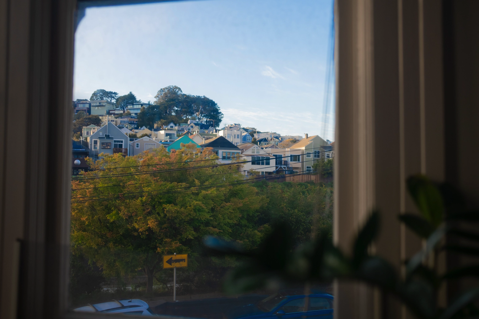 From a window, a string of houses huddle on a hillside. Trees. A yellow road sign pointing to the left.