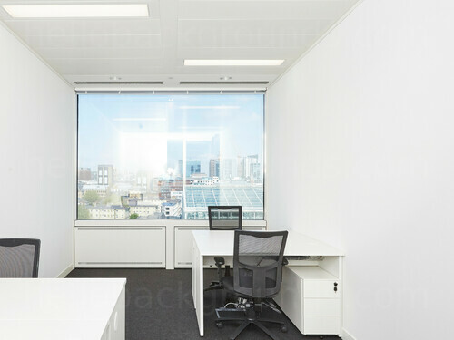 Compact shared workspace containing white desks with black mesh office chairs next to window with city view Google Meet background