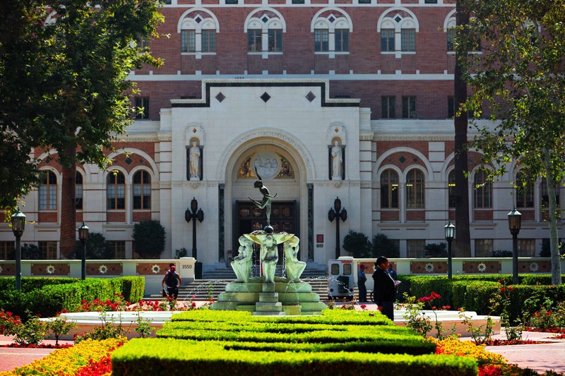 A USC campus building with a statue at the entrance and a fountain and gardens in the foreground