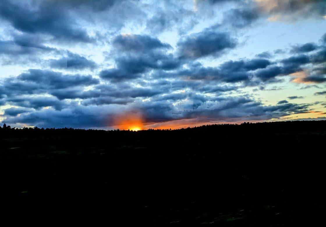Photo of a sunset. Pitch black landscape. A horizon line jagged by trees in a forest peeking up. The sky is light blue with a bunch of dark clouds. The top of the orange sun is barely visible above the horizon. It kinda looks like the forest is on fire.