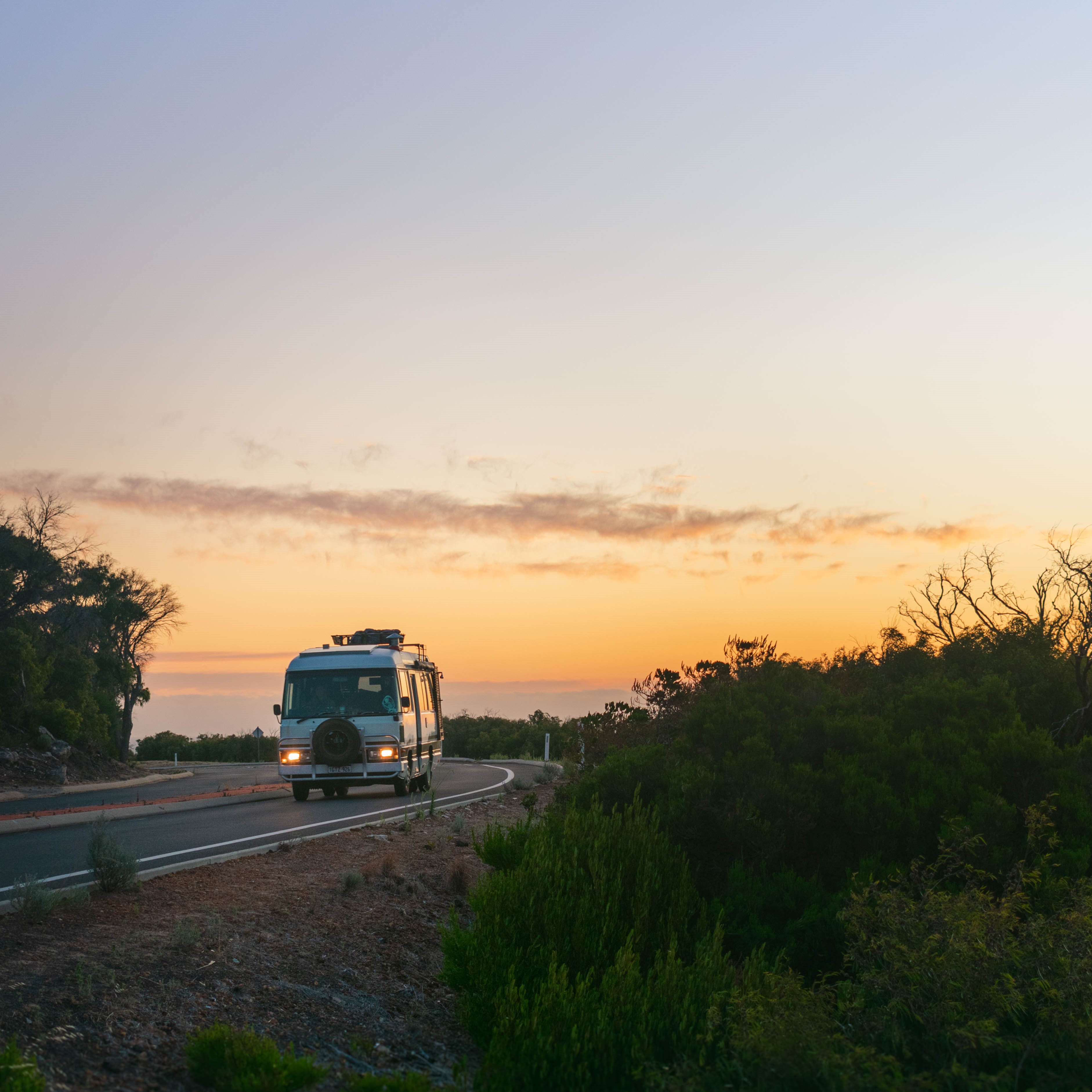 Landscape shot showing a road cutting through nature, the sun is setting and a van is in the middle of the road driving past.
