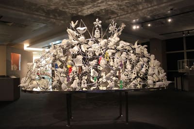 A large work sits on a metal table. It is a dome made up of smaller pieces of sculptures, strung together with wire.