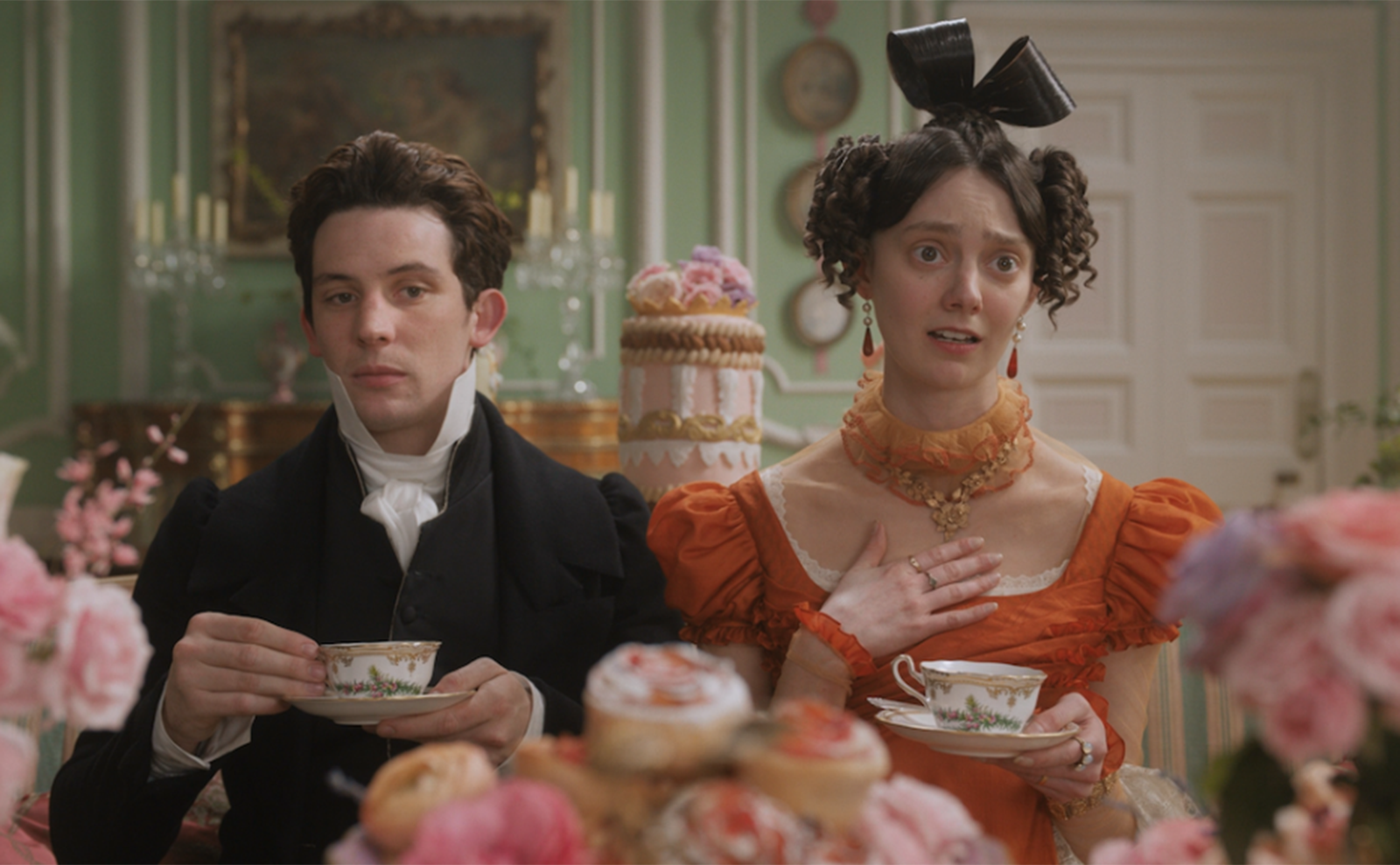 a young man and woman in regency costumes drinking tea with plates of cakes in the foreground