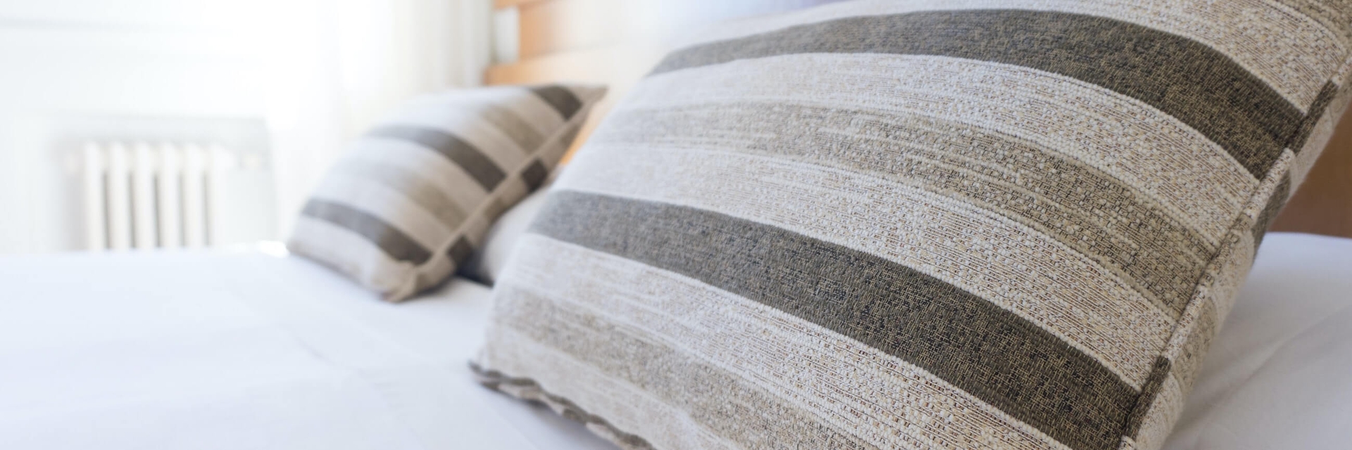 Travesseiros na cama de um hotel com lençol branco e fundo levemente borrado de uma janela com raios de sol.