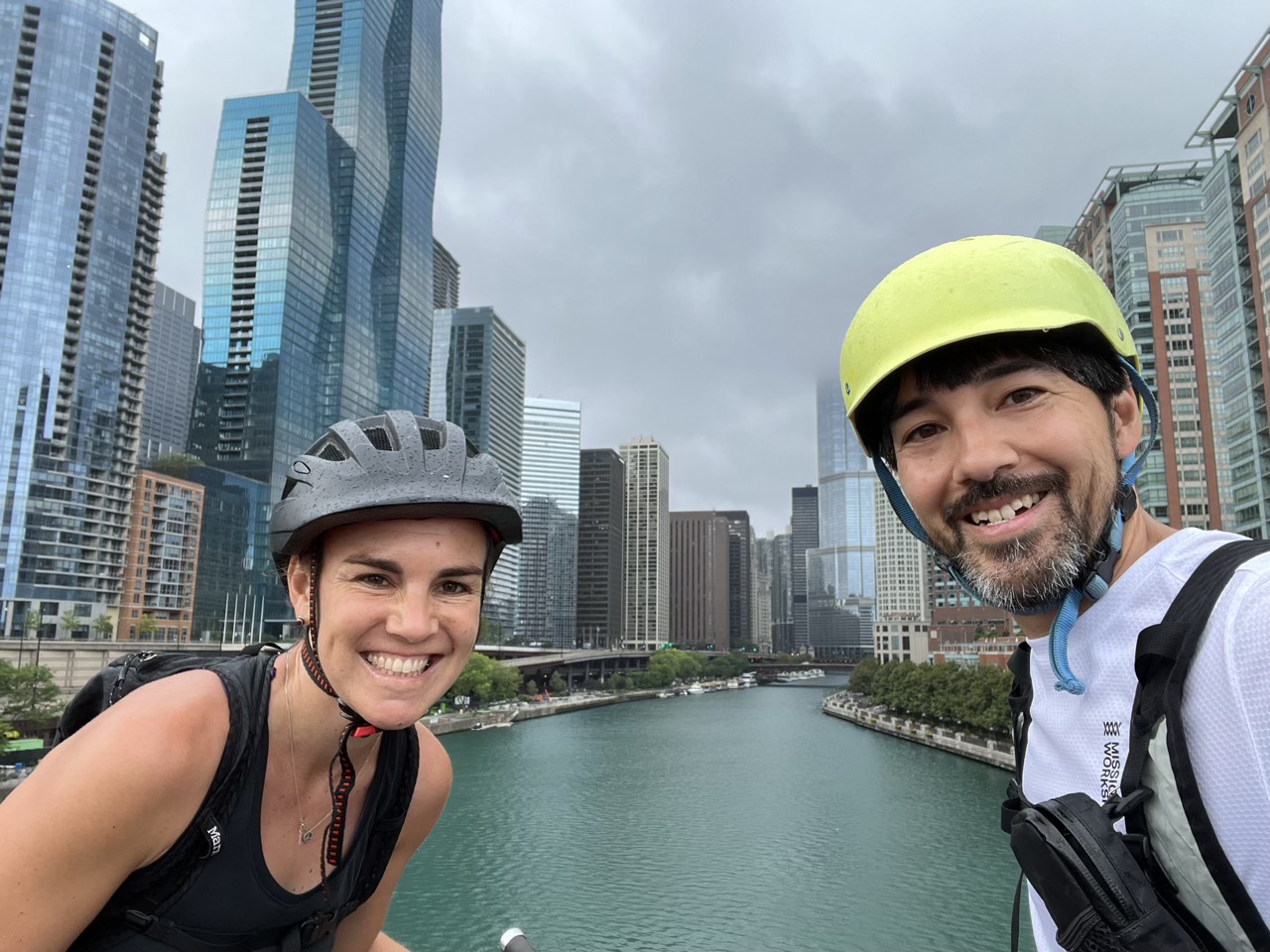 Cara and Benjamin on bikes with the Chicago skyline and river behind them