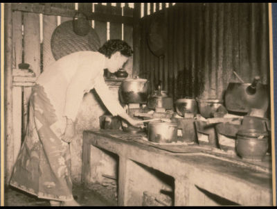 A housewife wearing a kebaya (a traditional Malay costume worn by women) bends over a concrete stove in a kampong kitchen.
