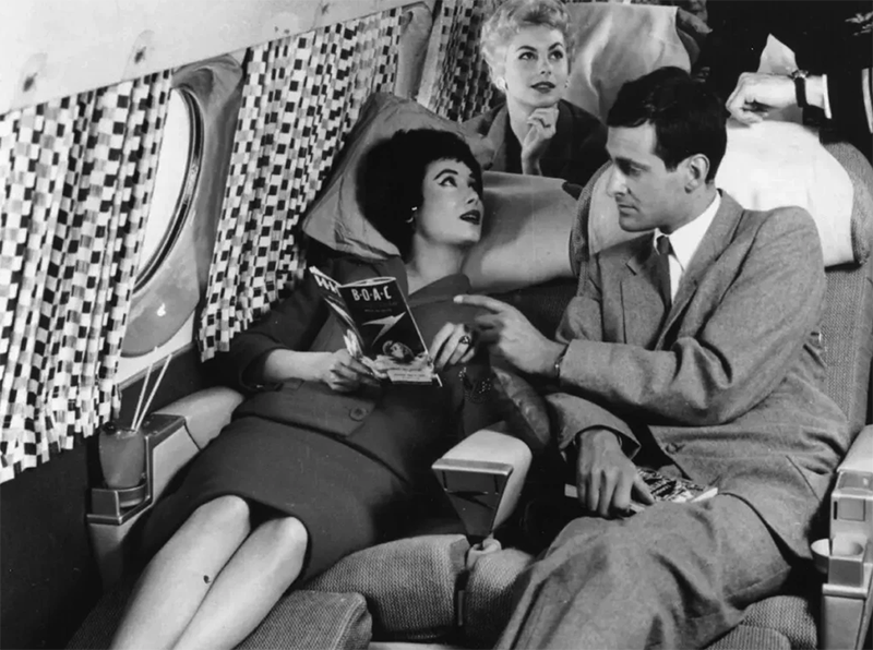  black and white photo of a woman and man on an airplane with the woman lying on a fold-out seat