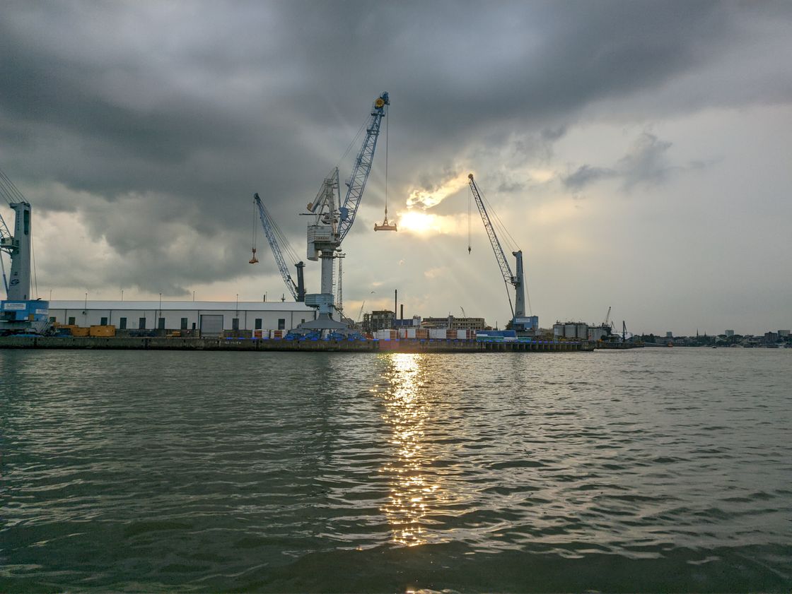 The setting sun is barely visible through a thick layer of dark clouds. The sunlight is reflected in the calm waves of a river. A few harbor cranes and small buildings reach into the dark sky.