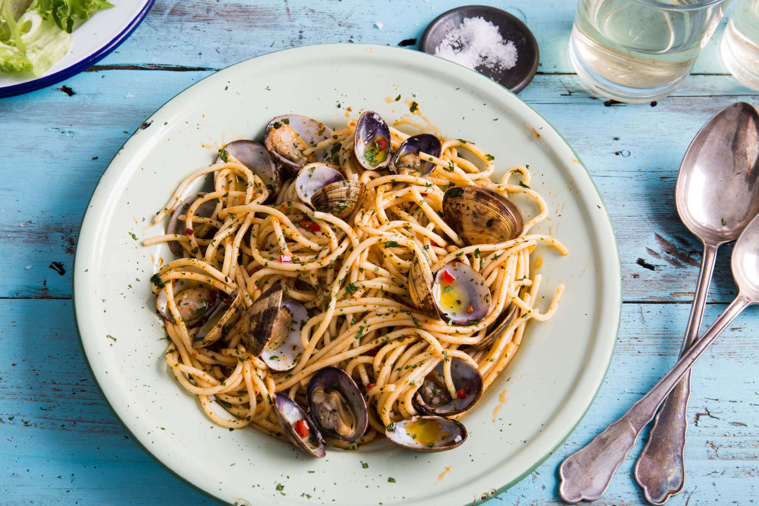 Clams ans spaghetti on a blue table with salad and wine glasses.