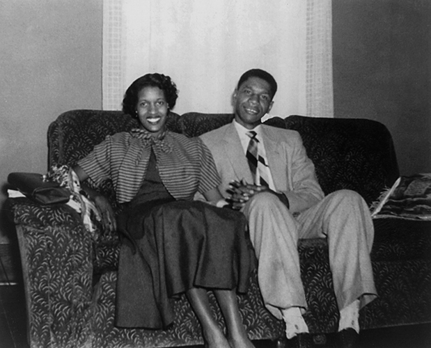 A black and white photo of Medgar and Myrlie Evers. They're seated on a couch, holding hands.