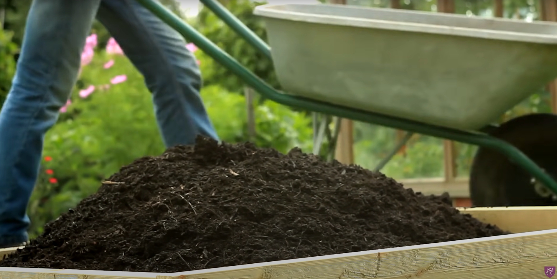 A raised bed with a pile of compost with a wheel barrow in the background
