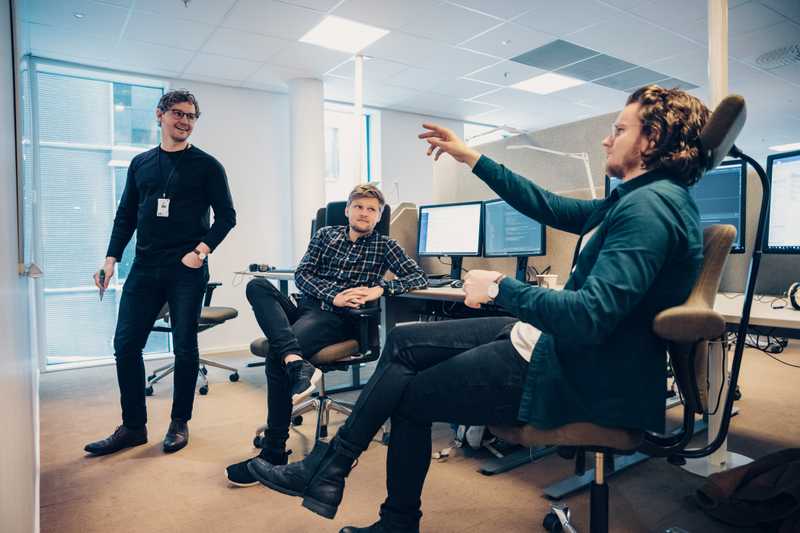 Tre lyshudede menn. En står og to sitterr på kontorstoler. En av dem som sitter peker på en whiteboard som er foran mennene.
