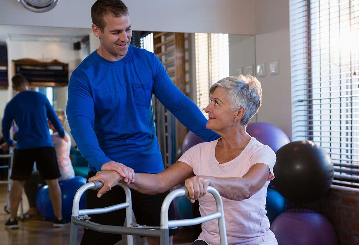 A man assists an elderly woman in an occupational therapy setting.