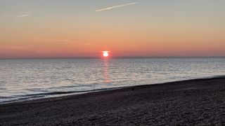 Sun setting over the sea with shingle in the foreground and the town of Worthing in the distance.
