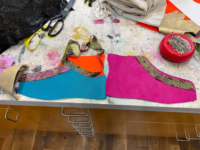 Top view of the work table
scattered with leather and fabric scraps,
scissors, and straight pins.
In the middle cut-out parts for one shoe --
magenta high-top outer,
with a floral fabric lacing panel,
back-to-back with a
cyan low-top inner with orange high-top upper,
and the same floral panel,
then a gold leather toe-cap.
