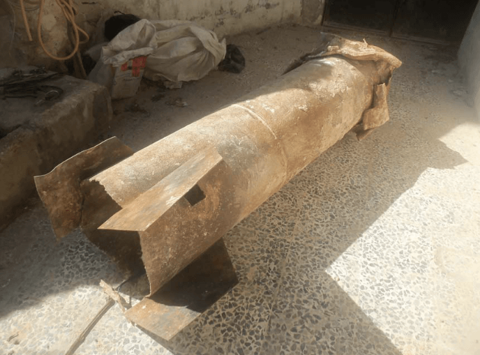 Photo shows a barrel  bomb laying on a tiled floor with metal fins welded on its lower body. The top of the barrel is crushed as a result of the drop impact. The barrel bomb was recovered after a chlorine attack in Hama in 2014. The photo appeared in a report by OPCW in 2014.