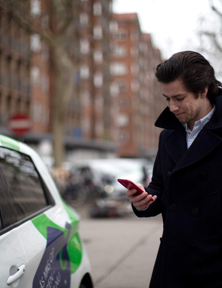 Green Mobility customer hero image featuring a caucasian man standing beside a car while looking down at his mobile device in Copenhagen.