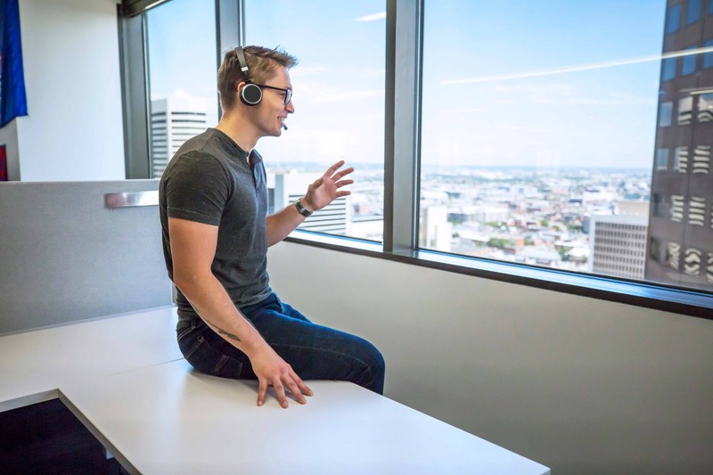 Man in casual clothing speaks on a wireless headset next to a window on an upper floor of an office building