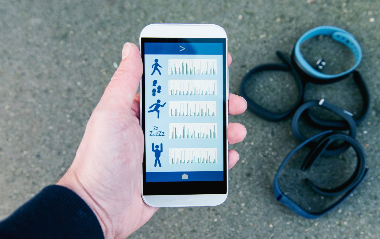 A white person's hand holding a phone with a wellness app interface visible on the screen, featuring running, walking, and sleeping icons, surrounded by various wearable workout bands on a concrete background.
