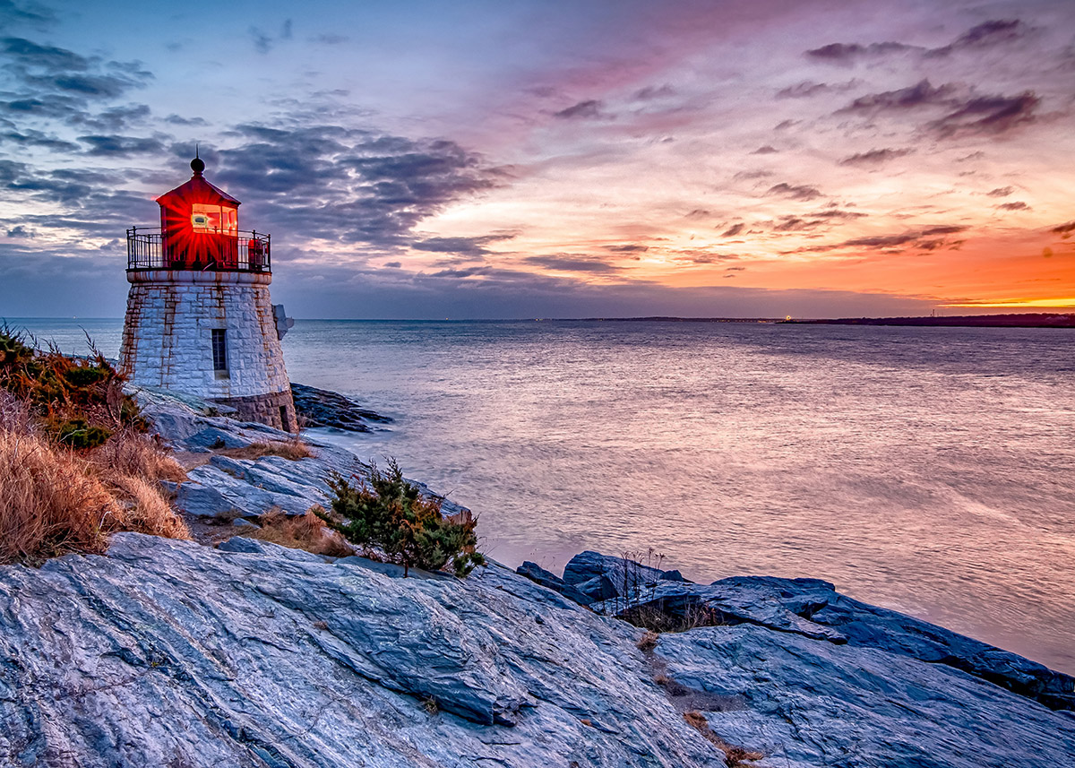 a white stone lighthouse on a rocky shore at dusk with the light shining on the ocean