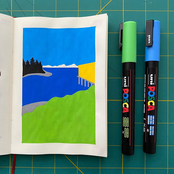 A blank notebook with a paint pen drawing of a bay with mountains behind it. A green and a blue pen are laid next to the notebook.