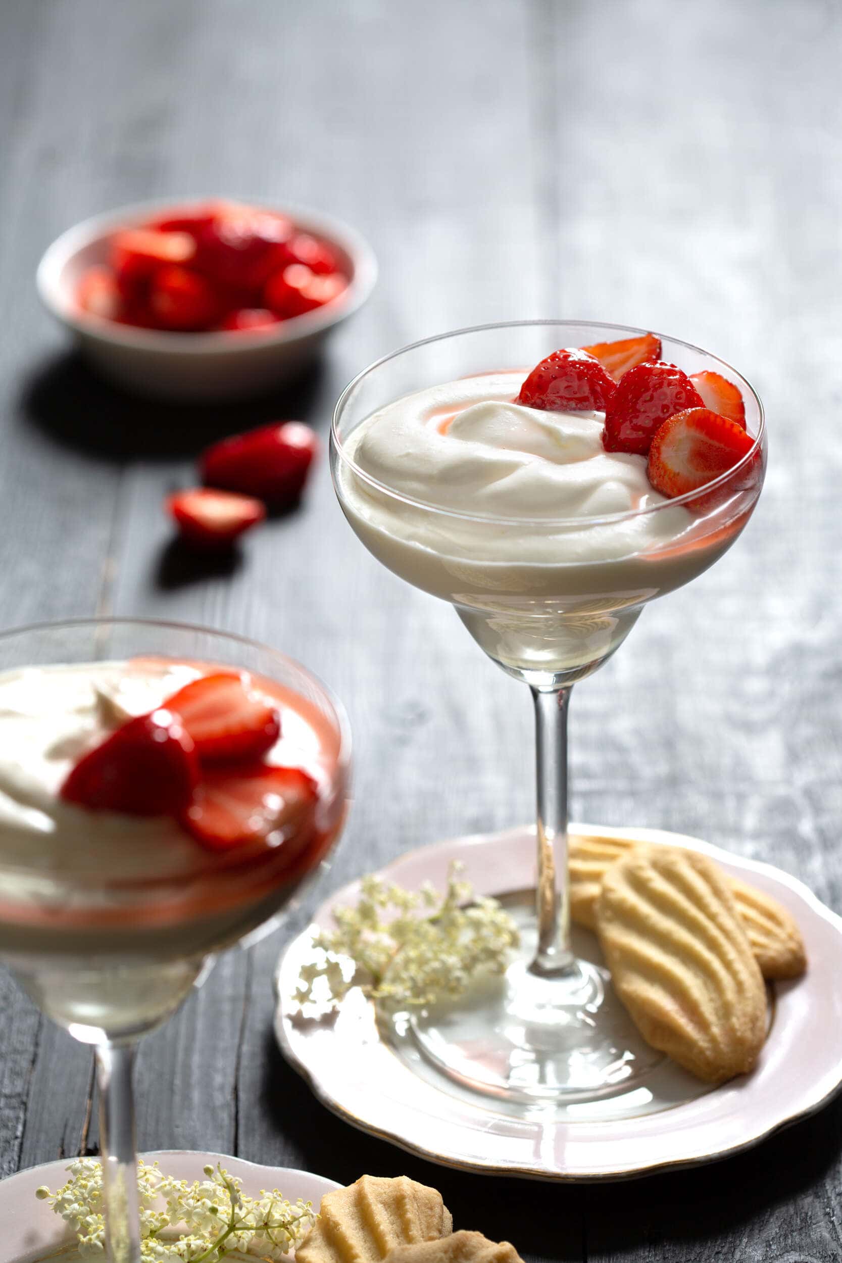 Elderflower Syllabub with Strawberries and biscuits set in a tall glass and strawberries in a bowl in the background.