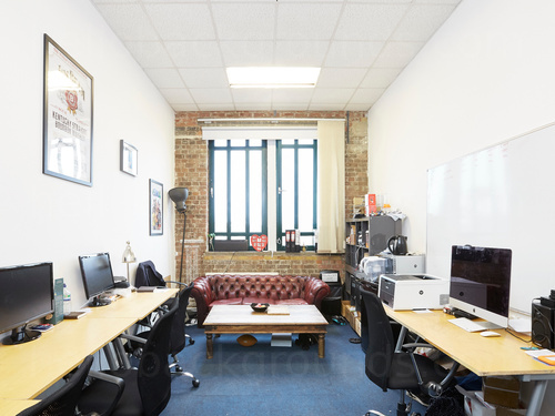 Compact office workspace lined with wooden computer desks with chairs and featuring red chesterfield sofa in front of window Google Meet background