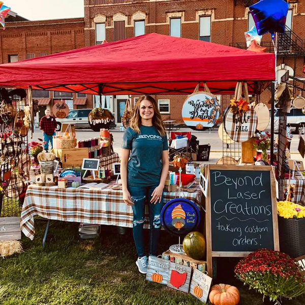Jena Stearns standing in front of an outdoor booth full of her custom products. There is a red awning over the booth, a plaid tablecloth covering the table, and lots of fall colors.