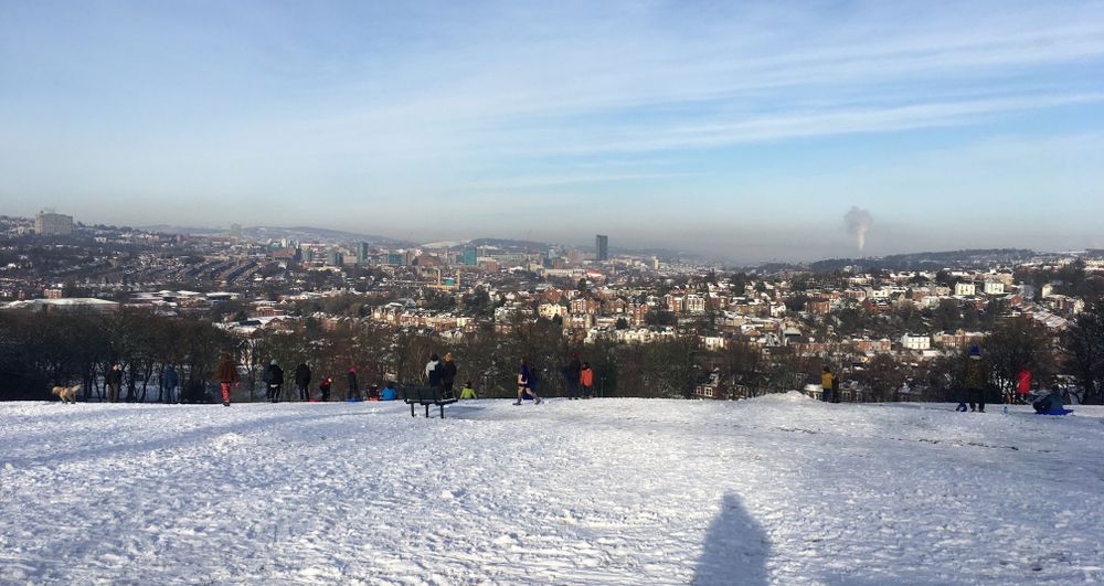 A snowy hill. People snowboard and ski. On the horizon trees a stunning view of the city nestled in the hills.