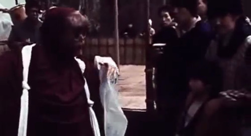 A screenshot from the horror film 'Noroi: The Curse' of a ritual featuring a dancer with a red kimono and disfigured mask.