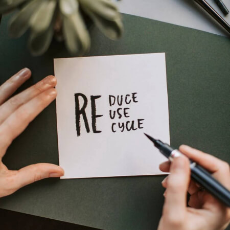 Zoomed in view of a hand writing 'Reduce, Reuse, Recycle' with a marker on a paper.